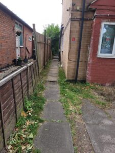 Property for sale A narrow concrete path runs between two brick buildings, with grass and plants along the edges—a potential investment property in Leicester. One building has a window, the other potted plants, and a wooden fence divides this split unit investment area. Presented by Palace Auctions