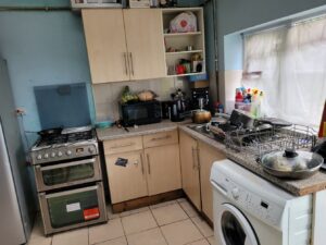 Property for sale A small kitchen with light wood cabinets anchors this Abbey Lane Investment Property, featuring a gas stove with a frying pan, microwave, dishes and utensils on drying racks, cleaning supplies by the window, and a washing machine in the foreground. Presented by Palace Auctions