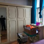 Property for sale A bedroom in an Abbey Lane Investment Property features cream-colored wardrobes, one with a mirrored door reflecting a person taking a photo. A box and plastic bin rest on a chair and desk by the window—perfect for 2-bed houses in Leicester. Presented by Palace Auctions