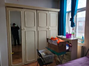 Property for sale A bedroom in an Abbey Lane Investment Property features cream-colored wardrobes, one with a mirrored door reflecting a person taking a photo. A box and plastic bin rest on a chair and desk by the window—perfect for 2-bed houses in Leicester. Presented by Palace Auctions