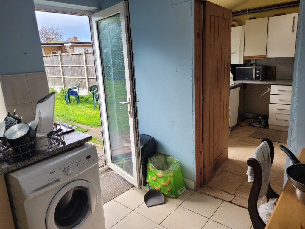 Property for sale A small kitchen in one of the 2-bed houses Leicester offers, with a washing machine, dishes drying on the counter, and a door open to a grassy backyard. White cabinets, microwave, and chairs are visible, plus a green recycling bag and broom near the door. Presented by Palace Auctions