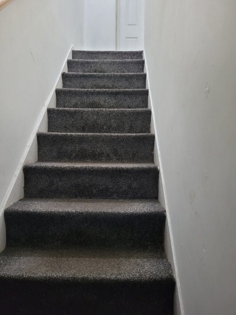 Property for sale A carpeted staircase with dark gray steps leads up between white walls to a closed white door at the top, creating an inviting entrance to this Abbey Lane investment property—an excellent split unit investment in Leicester. Presented by Palace Auctions
