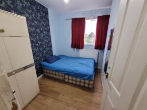 Property for sale A small bedroom with light blue walls, a bed with blue bedding, red curtains, wooden floor, patterned wallpaper on one wall, and a white wardrobe—ideal as an investment property Leicester buyers might find at a local property auction. Presented by Palace Auctions