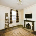 Property for sale A sparsely furnished room with bare wooden floors, a ladder against the wall, checked curtains, a wall-mounted TV above a black fireplace, and light walls with peeling paint—ideal for those seeking a Newhaven 2-bed maisonette investment. Presented by Palace Auctions