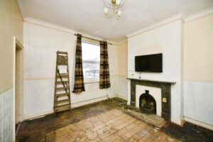 Property for sale A sparsely furnished room with bare wooden floors, a ladder against the wall, checked curtains, a wall-mounted TV above a black fireplace, and light walls with peeling paint—ideal for those seeking a Newhaven 2-bed maisonette investment. Presented by Palace Auctions