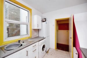Property for sale A small kitchen in this 2-bed maisonette Newhaven features white cabinets, a gray countertop, a window above the sink with a yellow frame, and modern appliances. A doorway with yellow trim opens to a red-walled area, highlighting this South Road investment. Presented by Palace Auctions