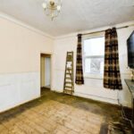 Property for sale An empty, unfurnished room in a Newhaven 2-bed maisonette investment, featuring bare wooden floors, cream walls, a wall-mounted TV, marble fireplace, large window with brown curtains, and a wooden ladder leaning against the wall. Presented by Palace Auctions
