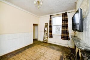 Property for sale An empty, unfurnished room in a Newhaven 2-bed maisonette investment, featuring bare wooden floors, cream walls, a wall-mounted TV, marble fireplace, large window with brown curtains, and a wooden ladder leaning against the wall. Presented by Palace Auctions