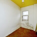 Property for sale A small, empty room in a 2-bed maisonette Newhaven, with stained white walls, yellow ceiling, brown floor, and mold near a window covered by a sheet. A radiator and large mirror lean against the right wall—ideal for investment or lease extension. Presented by Palace Auctions