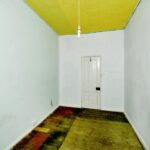 Property for sale A small, empty room with plain white walls, a yellow ceiling, and a single hanging lightbulb—part of a 2-bed maisonette Newhaven ideal for investment. A partially stripped wooden floor leads to a white door; a radiator sits on the right wall. Presented by Palace Auctions