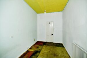 Property for sale A small, empty room with plain white walls, a yellow ceiling, and a single hanging lightbulb—part of a 2-bed maisonette Newhaven ideal for investment. A partially stripped wooden floor leads to a white door; a radiator sits on the right wall. Presented by Palace Auctions