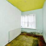 Property for sale A small, empty room with pale blue walls, a yellow ceiling, bare wooden floorboards, a radiator along one wall, and a window with sheer white curtains—mold visible near the window—offers renovation potential for a Newhaven 2-bed maisonette investment. Presented by Palace Auctions