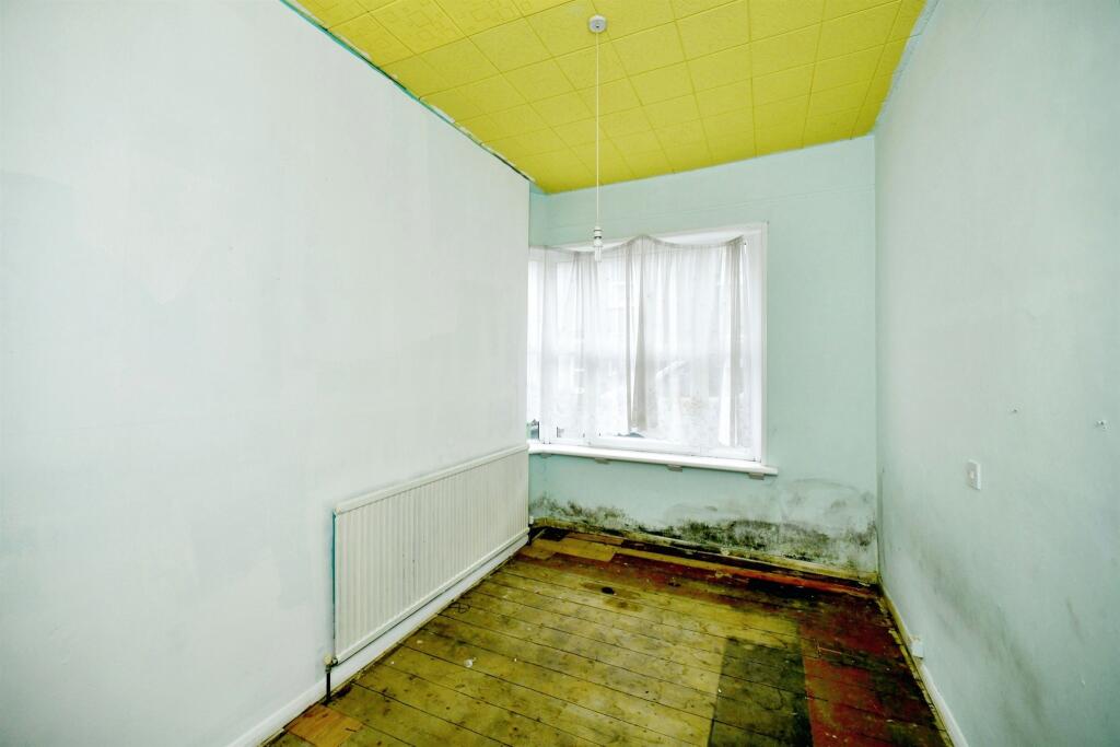 Property for sale A small, empty room with pale blue walls, a yellow ceiling, bare wooden floorboards, a radiator along one wall, and a window with sheer white curtains—mold visible near the window—offers renovation potential for a Newhaven 2-bed maisonette investment. Presented by Palace Auctions