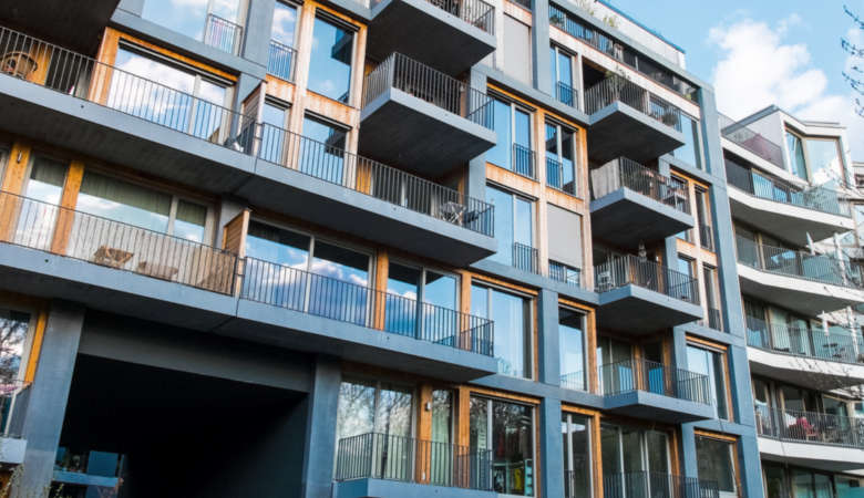 Property for sale Modern apartment building with large glass windows, wooden accents, and numerous balconies. The geometric design highlights contemporary features often seen in Leasehold Flats, popular in current UK architecture. Blue sky with some clouds above. Presented by Palace Auctions