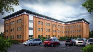 Property for sale A three-story office building with yellow brick walls and a green roof, soon to be a converted office apartment, is surrounded by a parking lot with several cars under a cloudy sky. Trees and bushes frame this Wath-Upon-Dearne apartment for sale. Presented by Palace Auctions