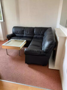 Property for sale A black leather L-shaped sofa is set in the corner of a Nottingham City rental property with beige walls and brown carpet, next to a small glass coffee table featuring a light-colored frame. Presented by Palace Auctions