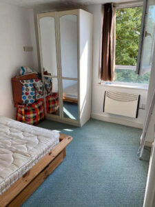 Property for sale A small, bright bedroom in a Nottingham City rental property, featuring a single bed frame and mattress, mirrored wardrobe, two bags on a colorful ottoman, blue carpet, and an open window with sunlight and views of green trees. Presented by Palace Auctions