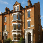 Property for sale A tall Victorian-style brick townhouse with large bay windows, white trim, ornate doorway, and a small front garden set against a blue sky—a classic London property investment opportunity. Presented by Palace Auctions