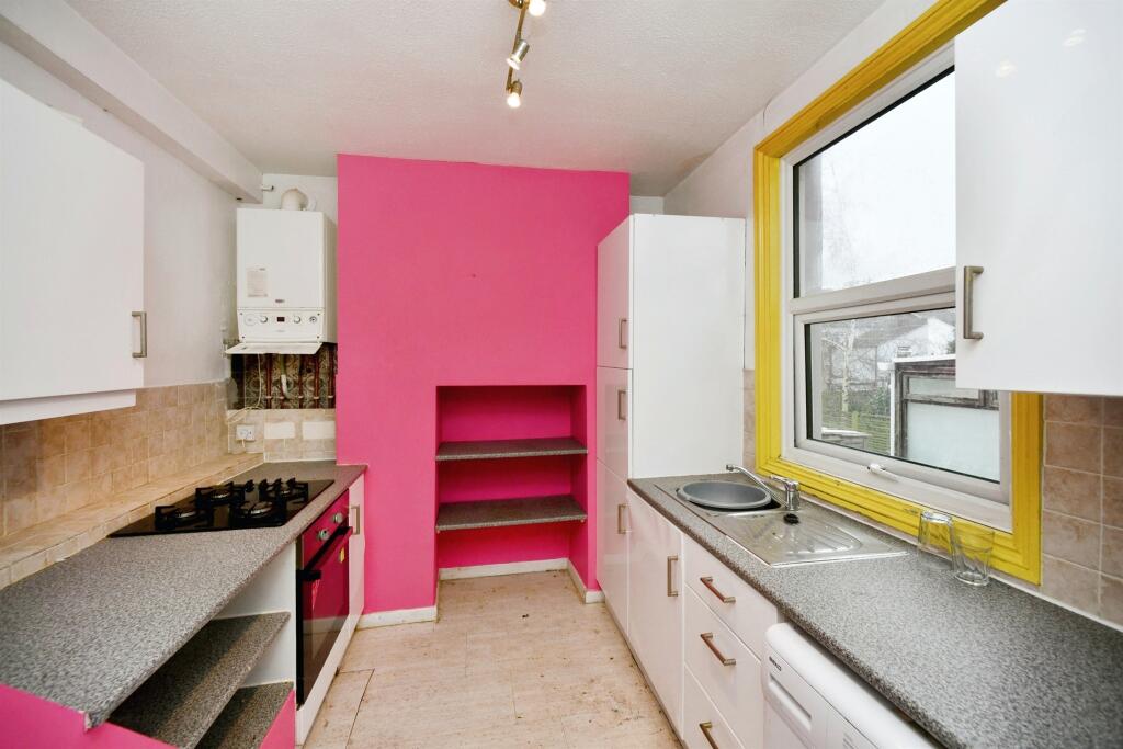 Property for sale A modern kitchen in a 2-bed maisonette Newhaven, featuring bright pink walls, white cabinets, grey countertops, a yellow window frame, built-in oven, gas stove, sink, and open shelves. Natural light floods through a large window. Presented by Palace Auctions