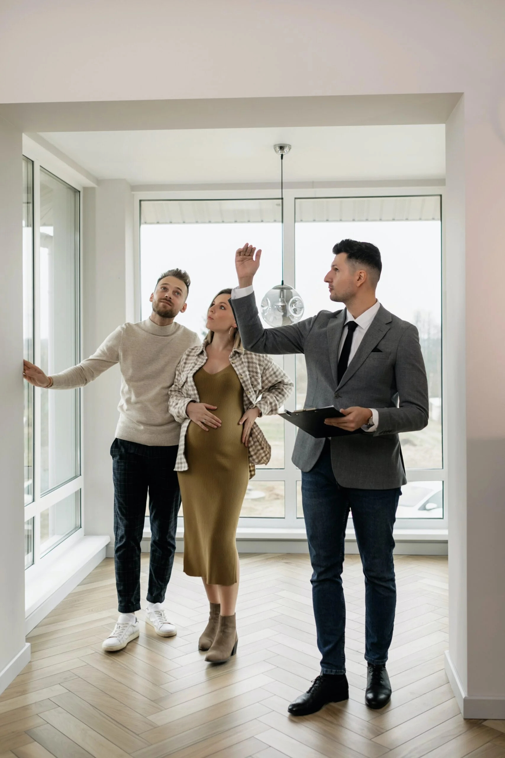 Property for sale A real estate agent, embodying why more real estate agents are becoming self-employed, shows a modern, bright apartment to a couple, including a pregnant woman. The agent gestures upwards with a tablet as the couple attentively looks around the room. Presented by Palace Auctions