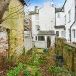 Property for sale Small, overgrown backyard with patchy grass, weeds, and mossy stone walls—ideal for a creative South Road investment. Surrounded by old white and brick houses; a bare tree and scattered items are visible. Presented by Palace Auctions