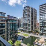 Property for sale Modern apartment buildings surround a canal with walkways, trees, and outdoor seating areas under a clear blue sky with a few clouds. The scene is viewed from a balcony above. Presented by Palace Auctions