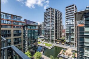 Property for sale Modern apartment buildings surround a canal with walkways, trees, and outdoor seating areas under a clear blue sky with a few clouds. The scene is viewed from a balcony above. Presented by Palace Auctions