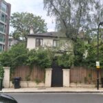 Property for sale A two-story Grade II listed London house with overgrown greenery covering the fence and part of the facade, set between a modern apartment building and tall trees, features trash bins by the sidewalk and a black gate in front. Presented by Palace Auctions