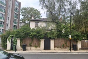 Property for sale A two-story Grade II listed London house with overgrown greenery covering the fence and part of the facade, set between a modern apartment building and tall trees, features trash bins by the sidewalk and a black gate in front. Presented by Palace Auctions