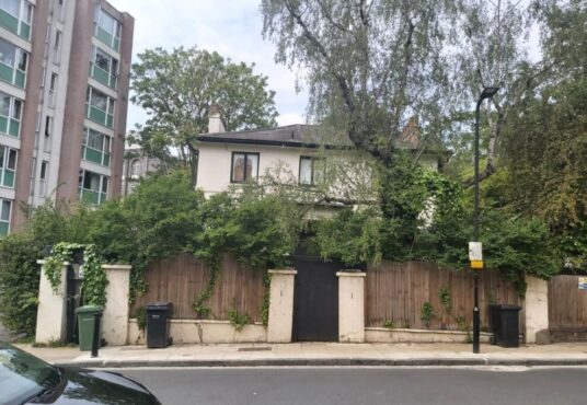 Property for sale A two-story Grade II listed London house with overgrown greenery covering the fence and part of the facade, set between a modern apartment building and tall trees, features trash bins by the sidewalk and a black gate in front. Presented by Palace Auctions