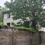 Property for sale A two-story white Grade II listed London house, partially hidden behind a tall brick wall and leafy tree, with other buildings, parked cars, and a sidewalk visible nearby on a cloudy day. Presented by Palace Auctions