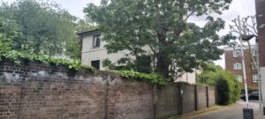 Property for sale A two-story white Grade II listed London house, partially hidden behind a tall brick wall and leafy tree, with other buildings, parked cars, and a sidewalk visible nearby on a cloudy day. Presented by Palace Auctions