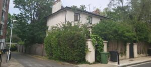 Property for sale A two-story house partially covered by green vines and trees stands at the corner of a quiet street on Greville Place NW6, with green and black bins by the entrance and a wooden fence running along the side. Presented by Palace Auctions