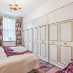 Property for sale A cozy bedroom in this prime central London flat features a double bed with white and red bedding, vintage wallpaper, built-in wardrobes with gold trim, sheer curtained window, and a charming vanity table with mirror. Presented by Palace Auctions