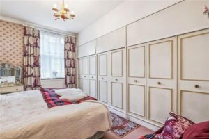 Property for sale A cozy bedroom in this prime central London flat features a double bed with white and red bedding, vintage wallpaper, built-in wardrobes with gold trim, sheer curtained window, and a charming vanity table with mirror. Presented by Palace Auctions