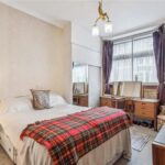 Property for sale A cozy bedroom in a Marylebone W1G flat for sale, featuring a double bed with a red plaid blanket, wooden dressers with mirrors, framed art, purple curtains, an armchair, and a vintage chandelier—ideal as an investment property in Marylebone. Presented by Palace Auctions