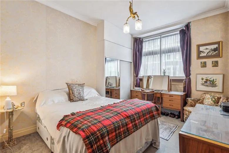 Property for sale A cozy bedroom in a Marylebone W1G flat for sale, featuring a double bed with a red plaid blanket, wooden dressers with mirrors, framed art, purple curtains, an armchair, and a vintage chandelier—ideal as an investment property in Marylebone. Presented by Palace Auctions