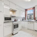 Property for sale A bright kitchen in a 3-bedroom apartment Marylebone features white cabinets, a stove, microwave, dishwasher, and a window with red curtains overlooking a stone building. Clear countertops hold just a kettle and a few small items. Presented by Palace Auctions