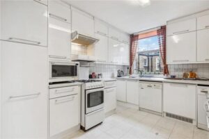 Property for sale A bright kitchen in a 3-bedroom apartment Marylebone features white cabinets, a stove, microwave, dishwasher, and a window with red curtains overlooking a stone building. Clear countertops hold just a kettle and a few small items. Presented by Palace Auctions