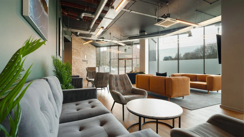 Property for sale A modern lounge at Langford Tower Sheffield features gray and tan sofas, armchairs, round tables, large windows, indoor plants, and industrial-style exposed ceilings—perfect for those seeking modern flats in Sheffield filled with natural light. Presented by Palace Auctions