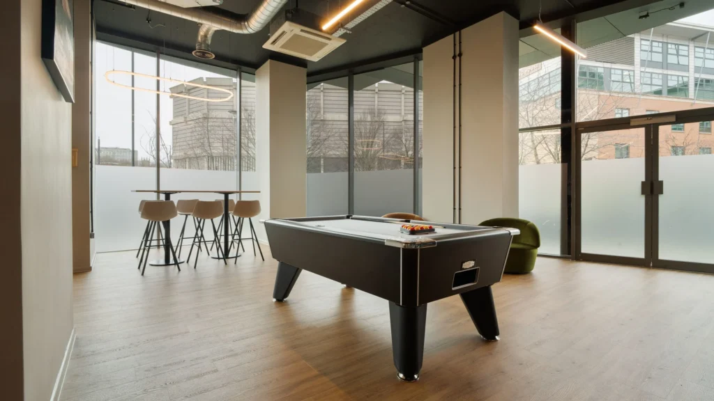 Property for sale Modern lounge in Langford Tower Sheffield, featuring a black pool table, green armchair, and tall table with barstools by large windows—perfect for those seeking modern flats in Sheffield. Presented by Palace Auctions