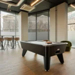 Property for sale Modern lounge in Langford Tower Sheffield, featuring a black pool table, green armchair, and tall table with barstools by large windows—perfect for those seeking modern flats in Sheffield. Presented by Palace Auctions