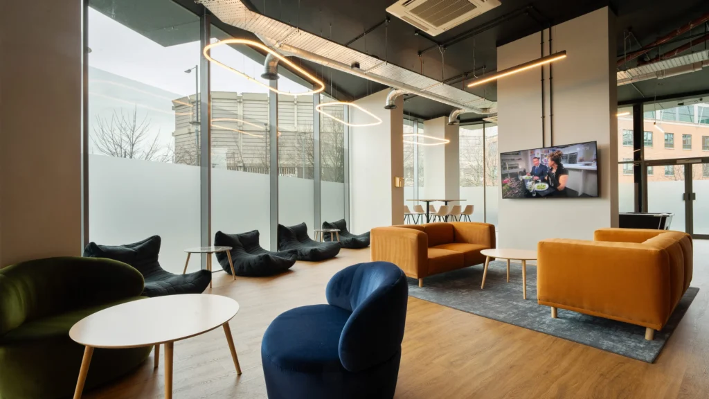 Property for sale Modern lounge with large windows, mustard and navy chairs, a TV on the wall, round coffee tables, and black bean bags along the window—perfect for those seeking stylish new build homes in Sheffield like Langford Tower Sheffield. Presented by Palace Auctions