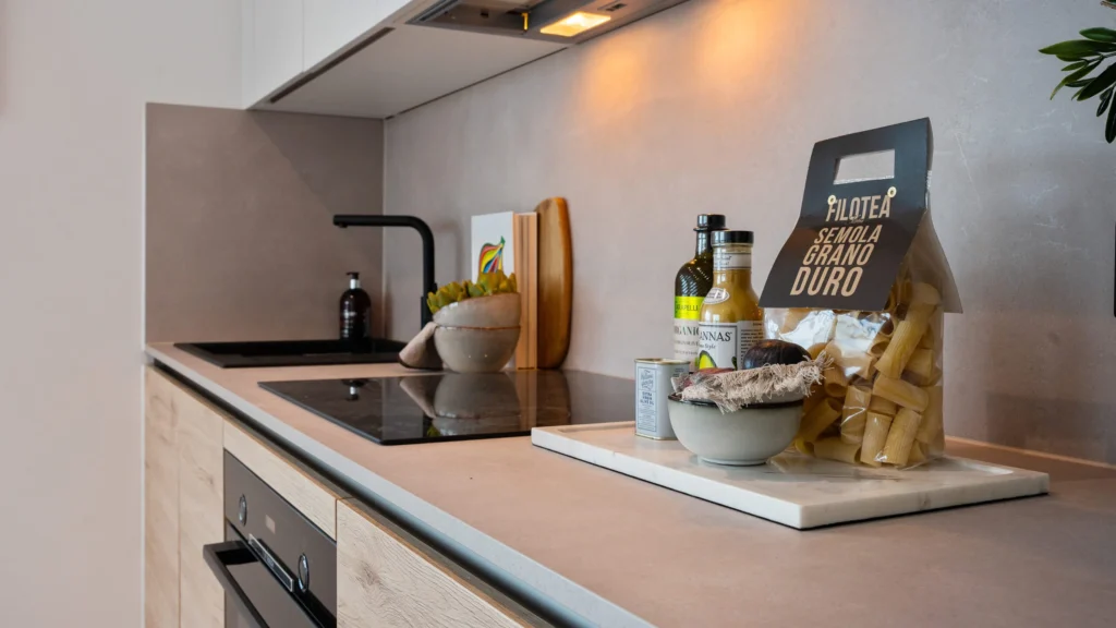 Property for sale A modern kitchen countertop, perfect for modern flats in Sheffield, with an electric stove, sink, olive oil, pasta, salt, a bowl, and a small potted plant on a white tray. Light wood cabinets add to the minimal decor. Presented by Palace Auctions