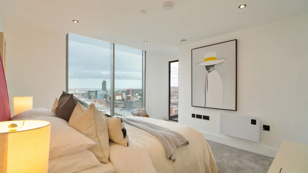 Property for sale A modern bedroom in Langford Tower Sheffield features large windows with city skyline views, a neatly made bed with neutral-toned pillows, and stylish framed artwork—ideal for those seeking apartments for sale in Sheffield. Presented by Palace Auctions