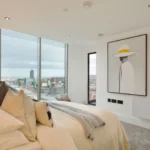 Property for sale A modern bedroom in Langford Tower Sheffield features large windows with city skyline views, a neatly made bed with neutral-toned pillows, and stylish framed artwork—ideal for those seeking apartments for sale in Sheffield. Presented by Palace Auctions