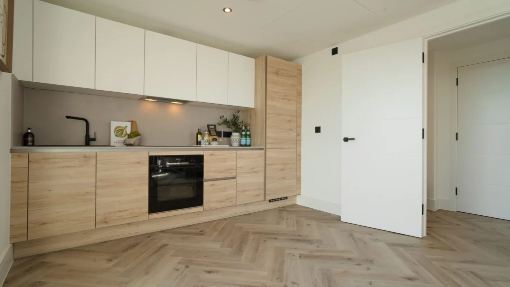 Property for sale Modern kitchen with light wood cabinets, white upper cupboards, a black oven, and a black faucet—perfect for modern flats in Sheffield. The herringbone floor and open white door add style; bottles and a plant decorate the countertop. Presented by Palace Auctions