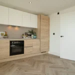 Property for sale Modern kitchen with light wood cabinets, white upper cupboards, a black oven, and a black faucet—perfect for modern flats in Sheffield. The herringbone floor and open white door add style; bottles and a plant decorate the countertop. Presented by Palace Auctions