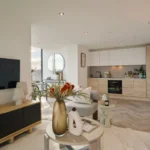 Property for sale Modern open-plan living room and kitchen in Langford Tower Sheffield, featuring light wood floors, neutral furniture, a TV on a wooden console, decorative vases on a round coffee table, and large windows flooding the space with natural light. Presented by Palace Auctions