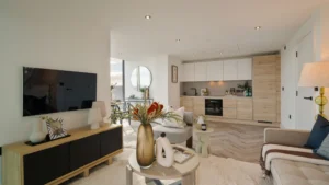 Property for sale Modern open-plan living room and kitchen in Langford Tower Sheffield, featuring light wood floors, neutral furniture, a TV on a wooden console, decorative vases on a round coffee table, and large windows flooding the space with natural light. Presented by Palace Auctions